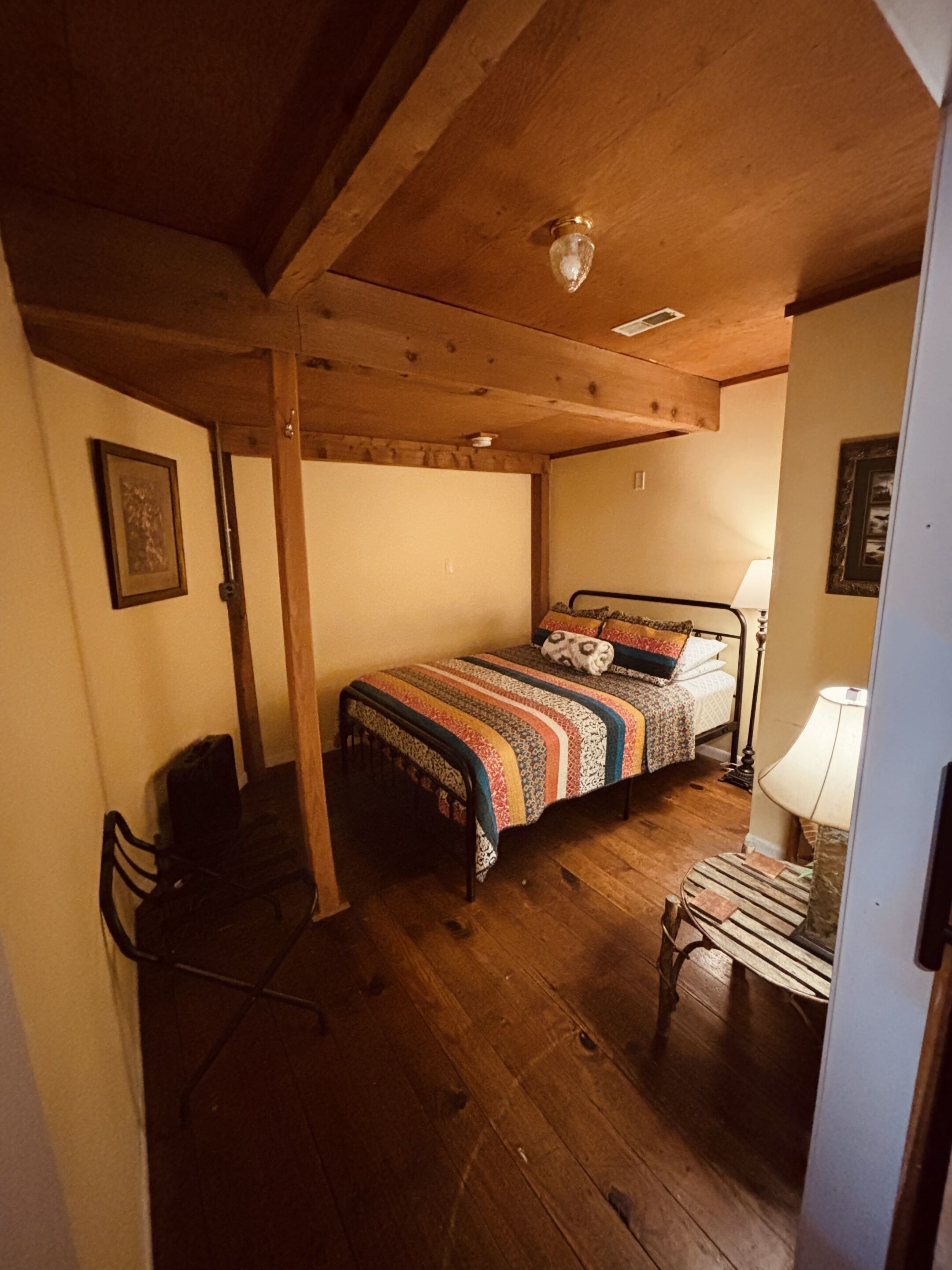A bedroom with a wooden ceiling, a bed with a patterned blanket, a bedside table with a lamp, and a chair.