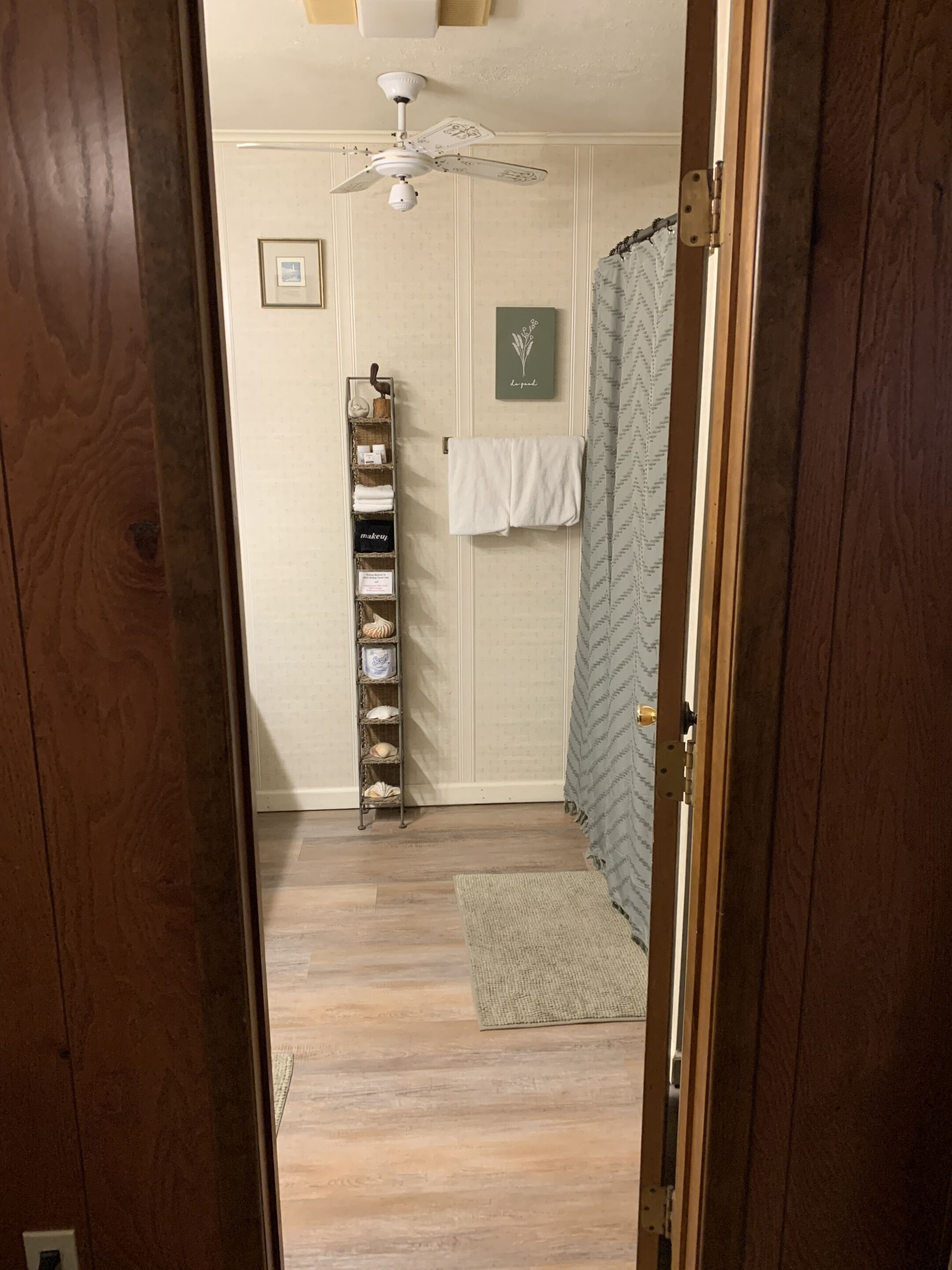 A bathroom with a wooden floor, a white ceiling fan, a towel rack, and a shower curtain.