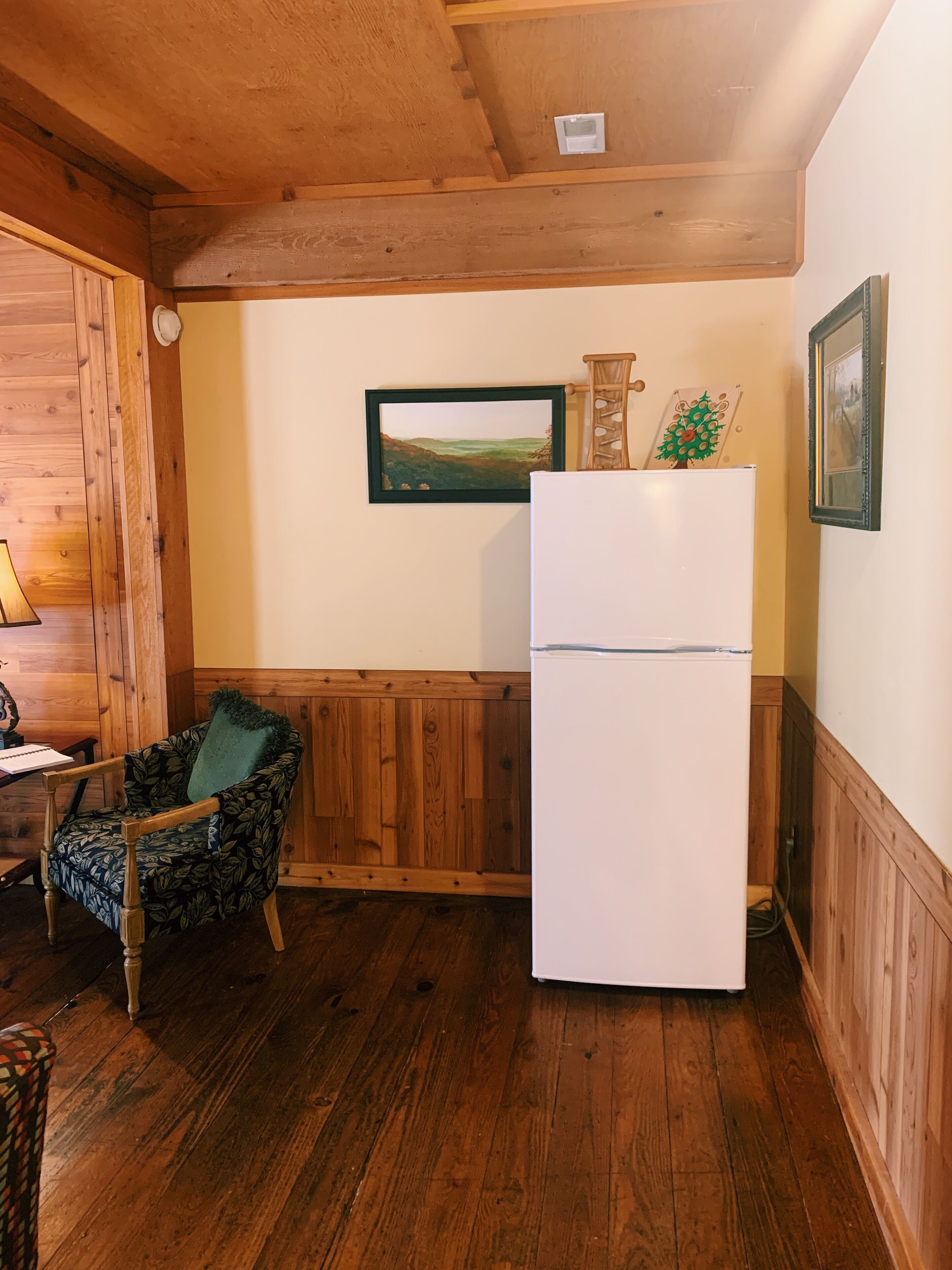Une petite pièce chaleureuse aux murs et au plafond en bois. Un réfrigérateur blanc est adossé au mur, et une chaise avec un coussin vert se trouve dans un coin.