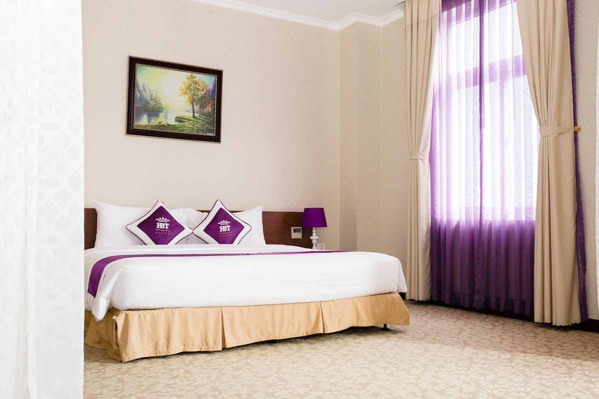 A hotel room with a large bed, a nightstand, a lamp, a painting on the wall, and a window with curtains.