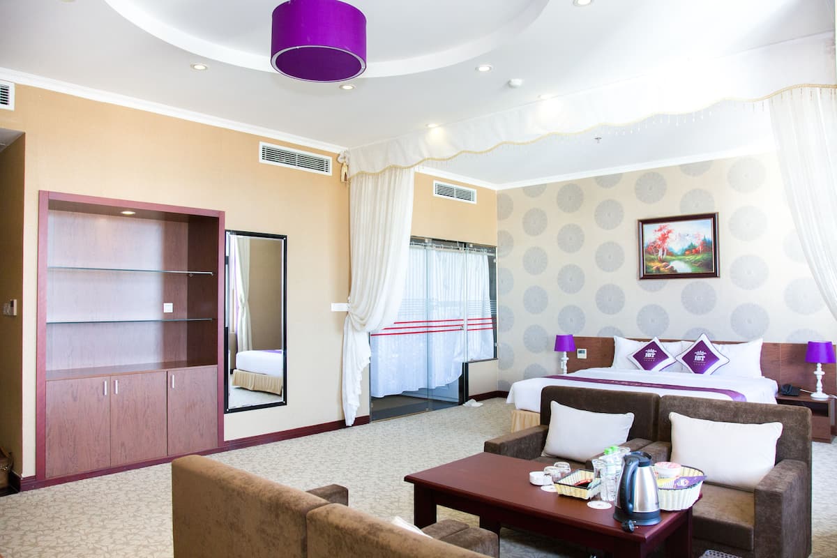 A hotel room with a bed, a sofa, a coffee table, a wardrobe, and a bathroom visible through a glass door.