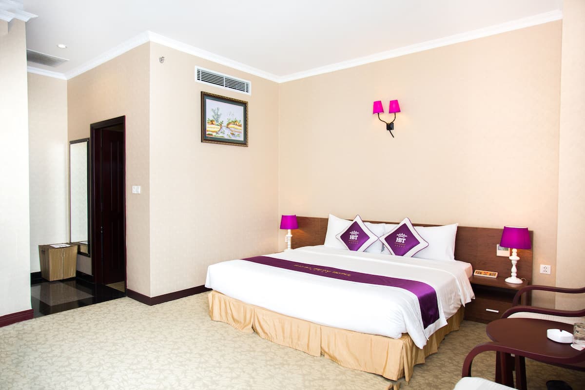 A hotel room with a large bed, two bedside tables, a small desk, a chair, a waste basket, and a picture on the wall.