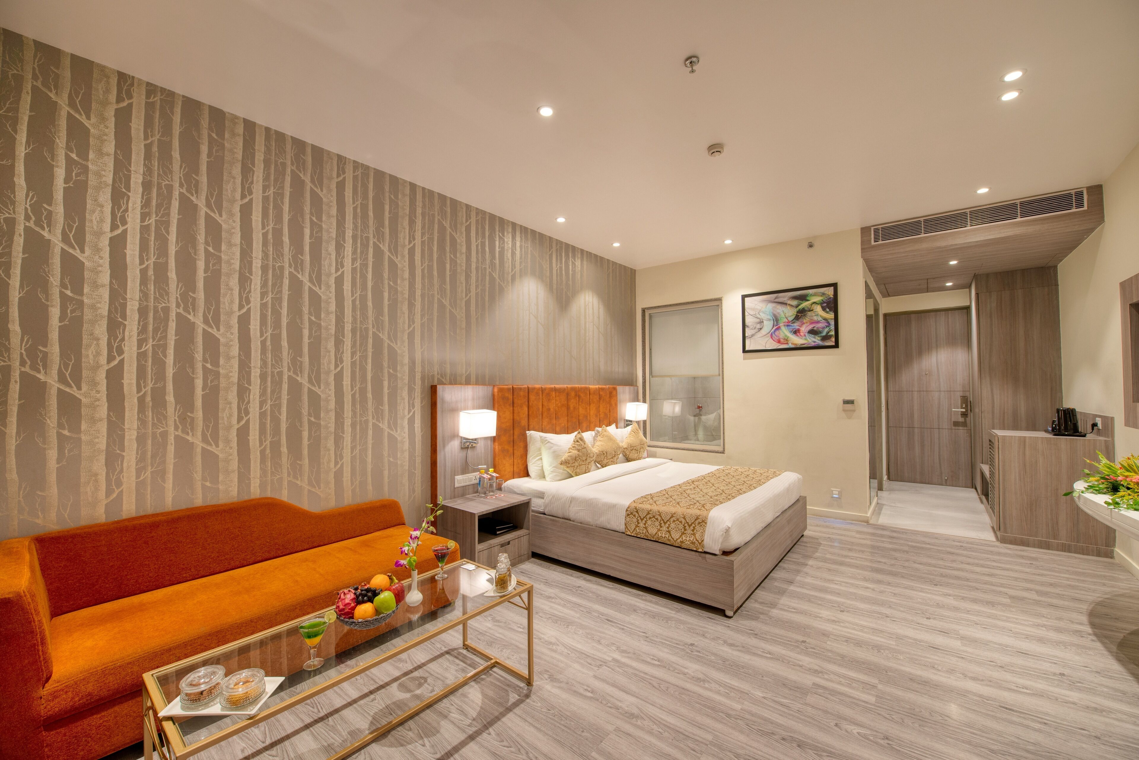 A hotel room with a bed, a sofa, a nightstand with a lamp, a small table with a tray of items, and a bathroom visible in the background.