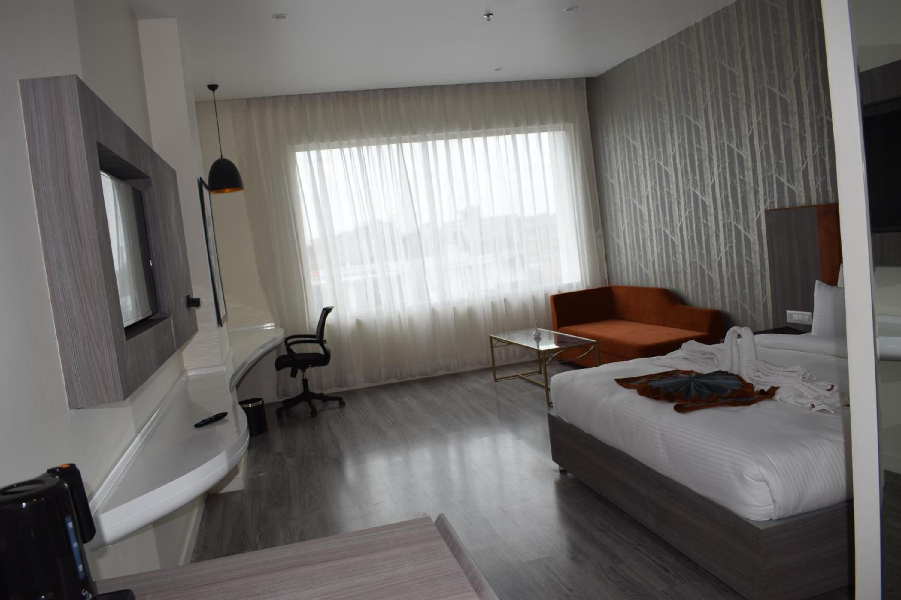 A modern hotel room with a bed, a desk, a chair, a small table, and a large window with sheer curtains.