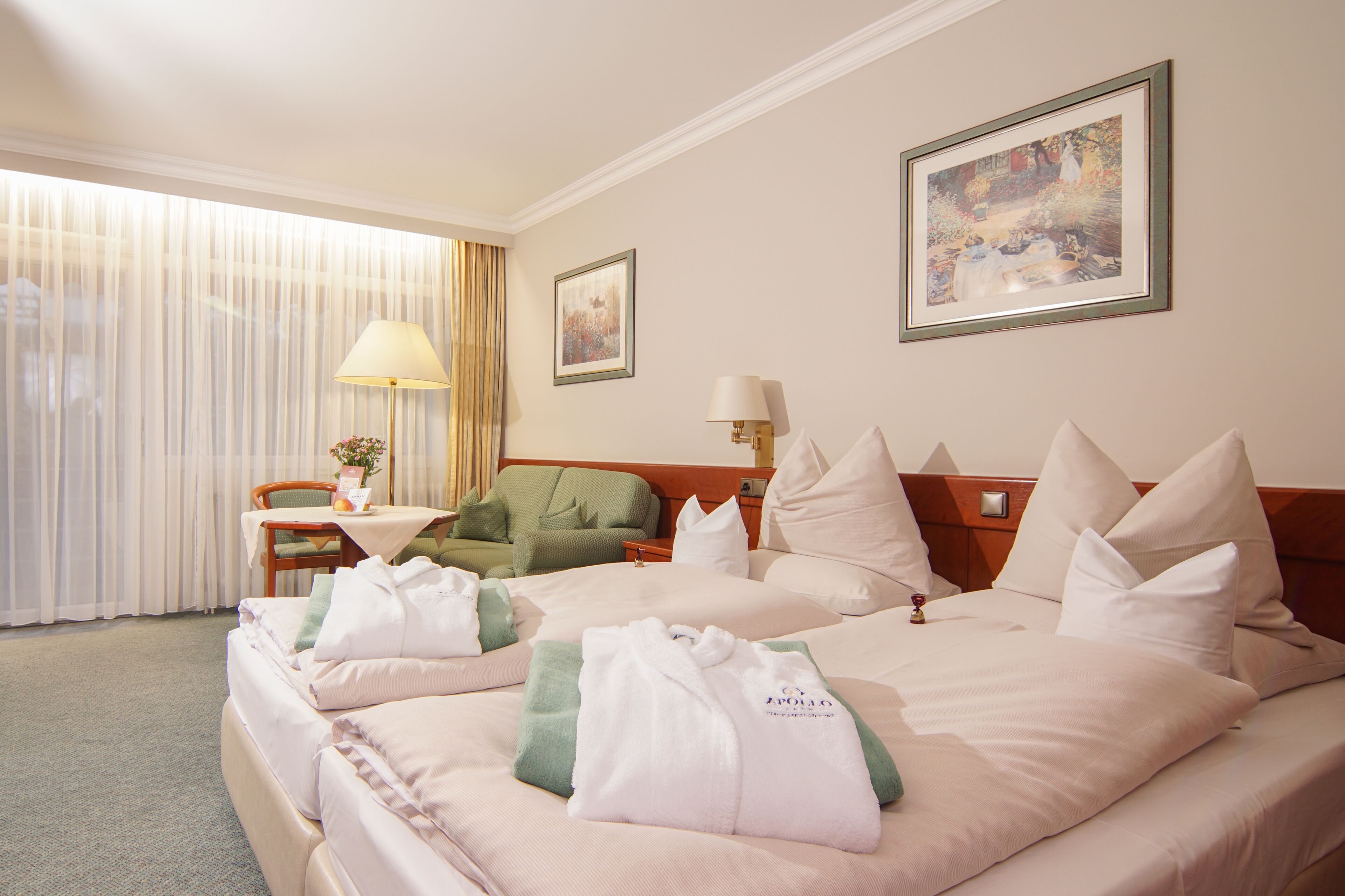 A hotel room with two beds, a sitting area with a table and chair, and a large window with curtains.