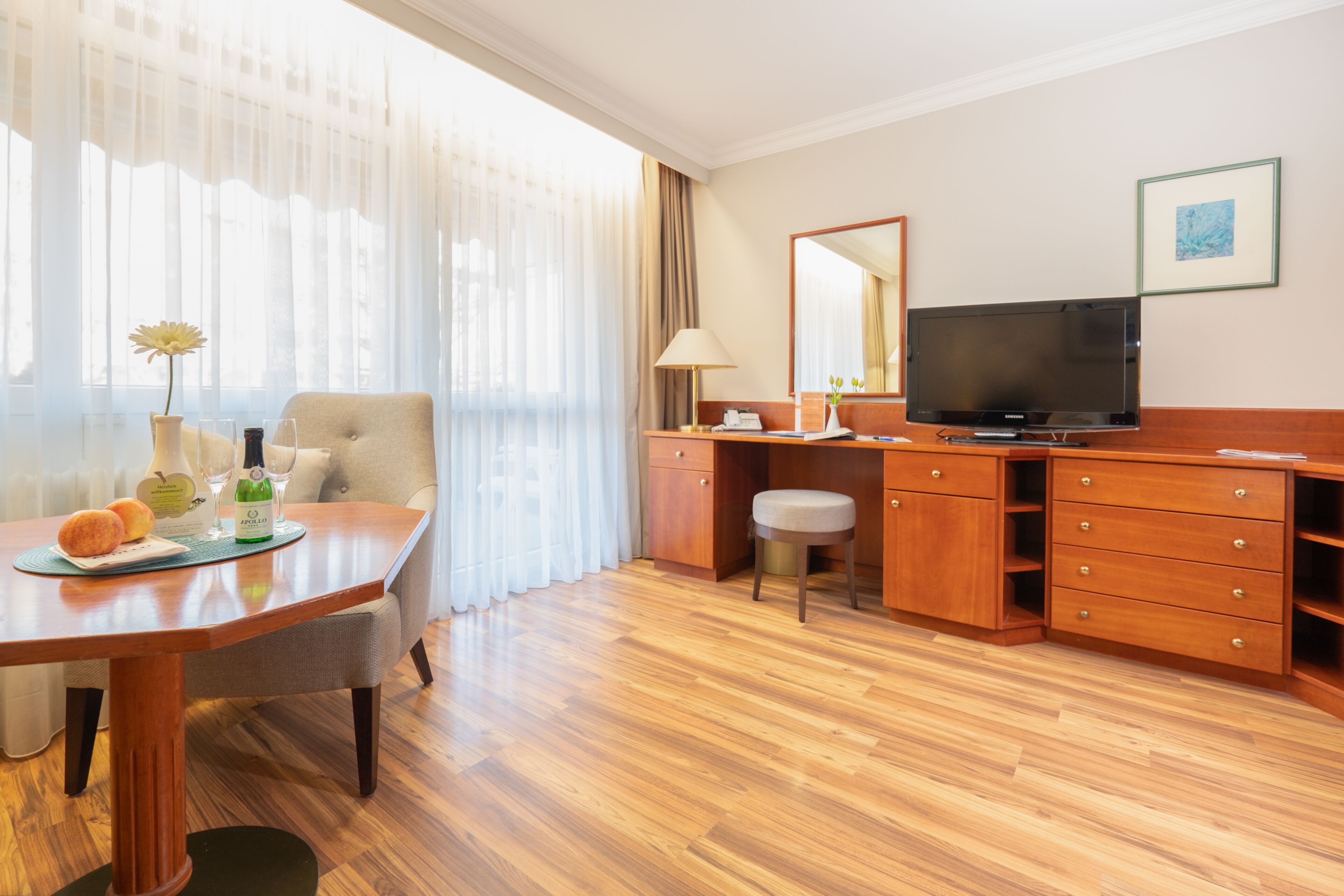 A hotel room with a wooden floor, a dining table with a vase of flowers, a television, and a desk with a lamp.