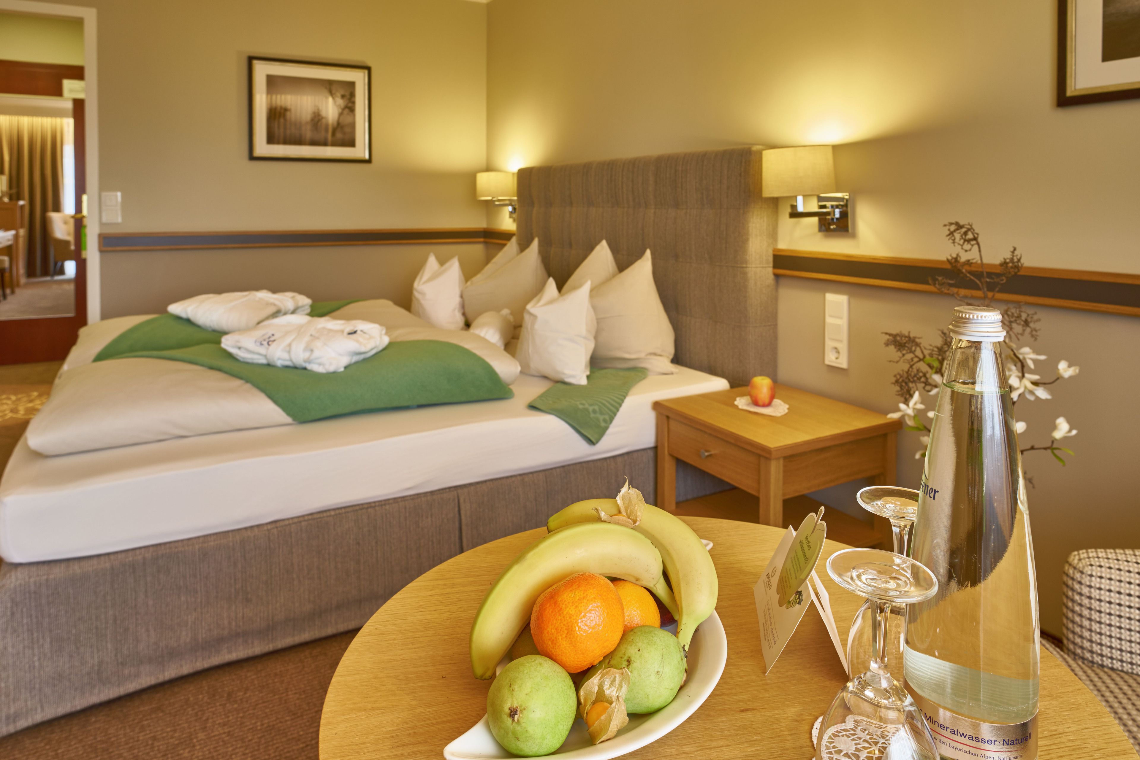 Habitación de hotel con una cama bien hecha, una mesita de noche con una botella y vasos, y un frutero sobre la mesa.