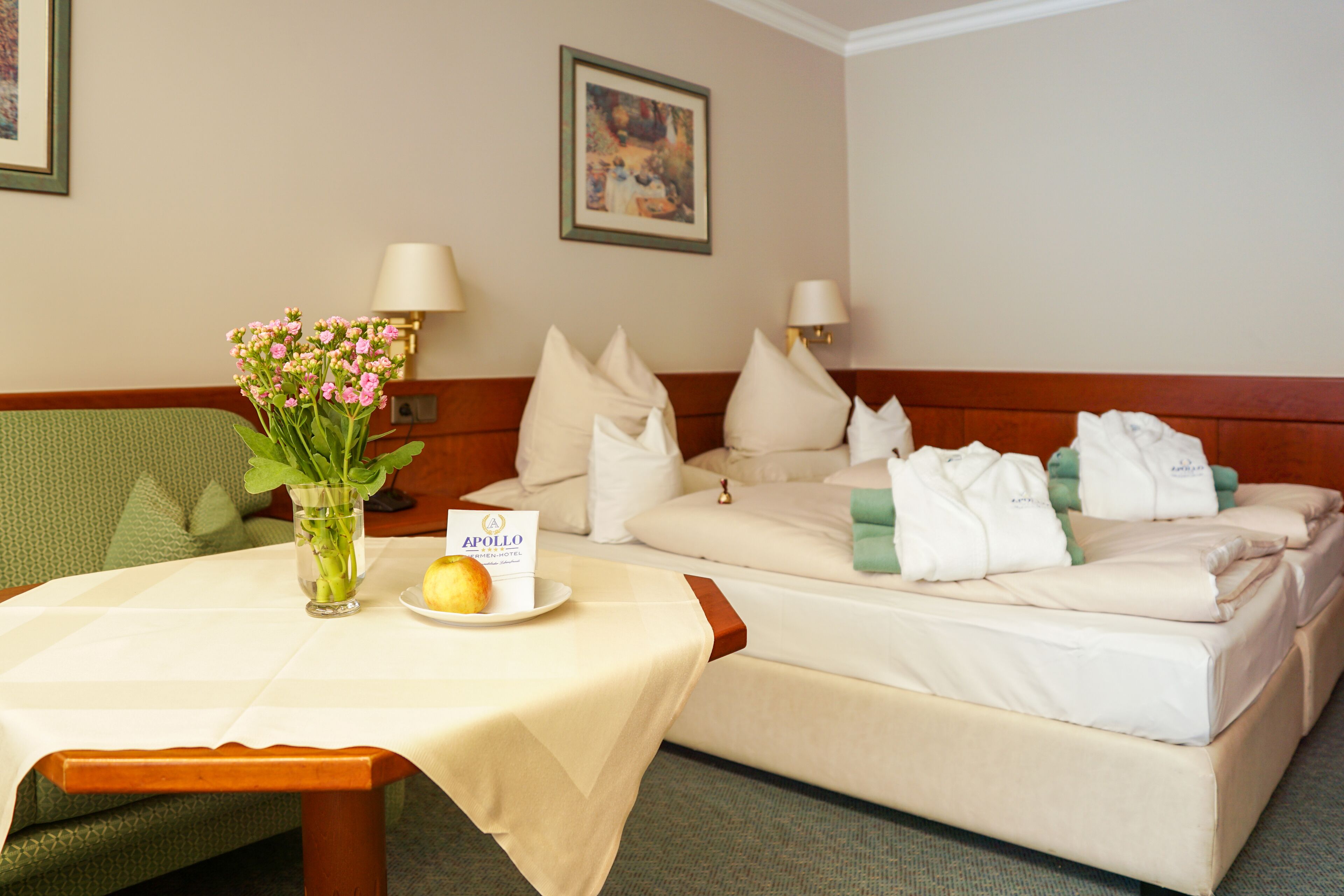Una habitación de hotel con una cama, una mesa con un jarrón de flores, un plato de fruta y un cartel.