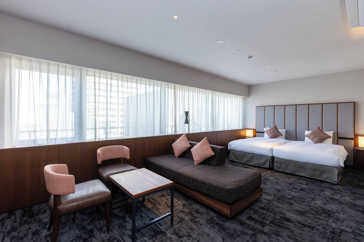 Ein modernes Hotelzimmer mit einer Couch, zwei Betten, einem Esstisch und einem großen Fenster mit Vorhängen.