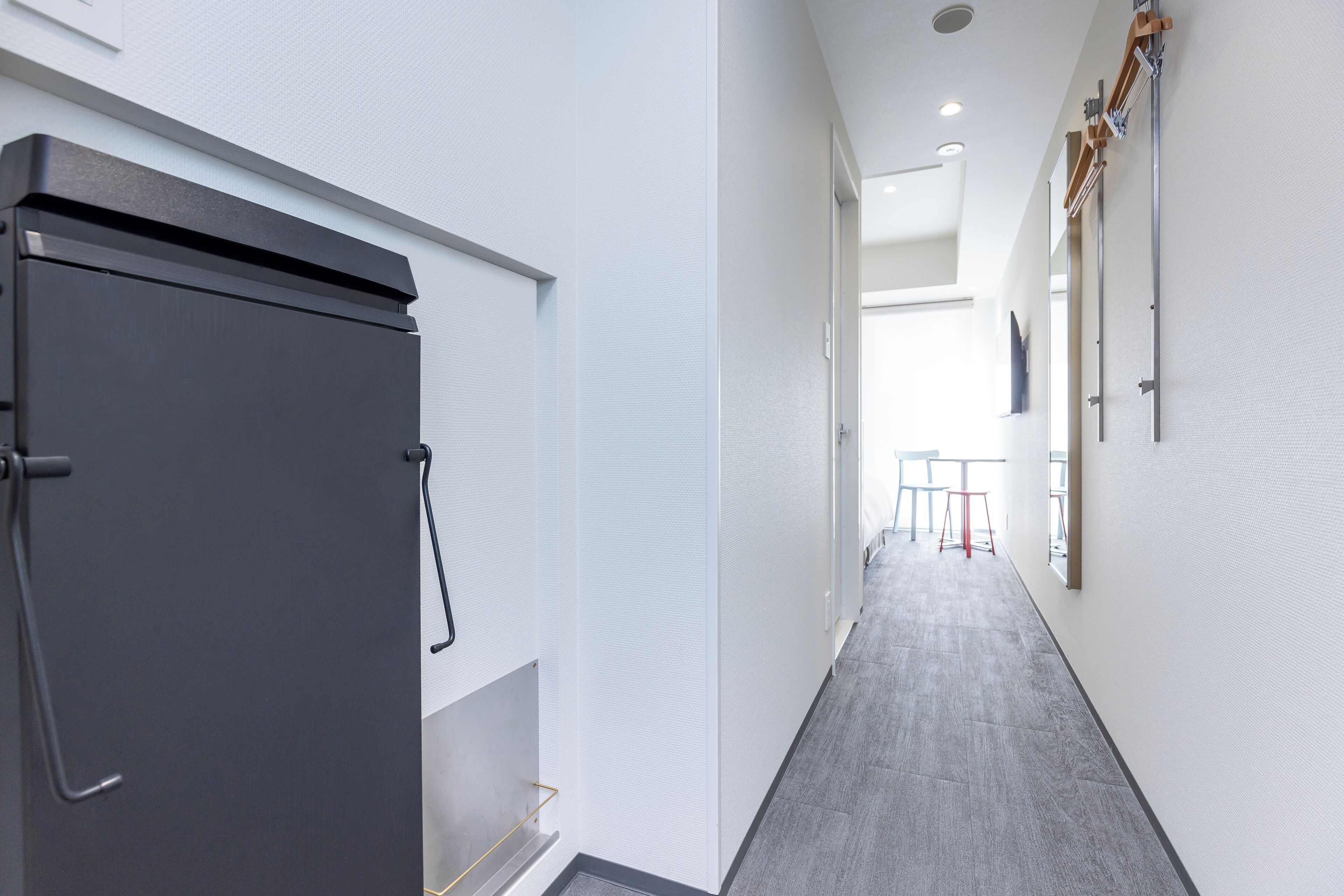 A hallway with a grey door, white walls, and a grey carpet. There are two tables and chairs in the distance.