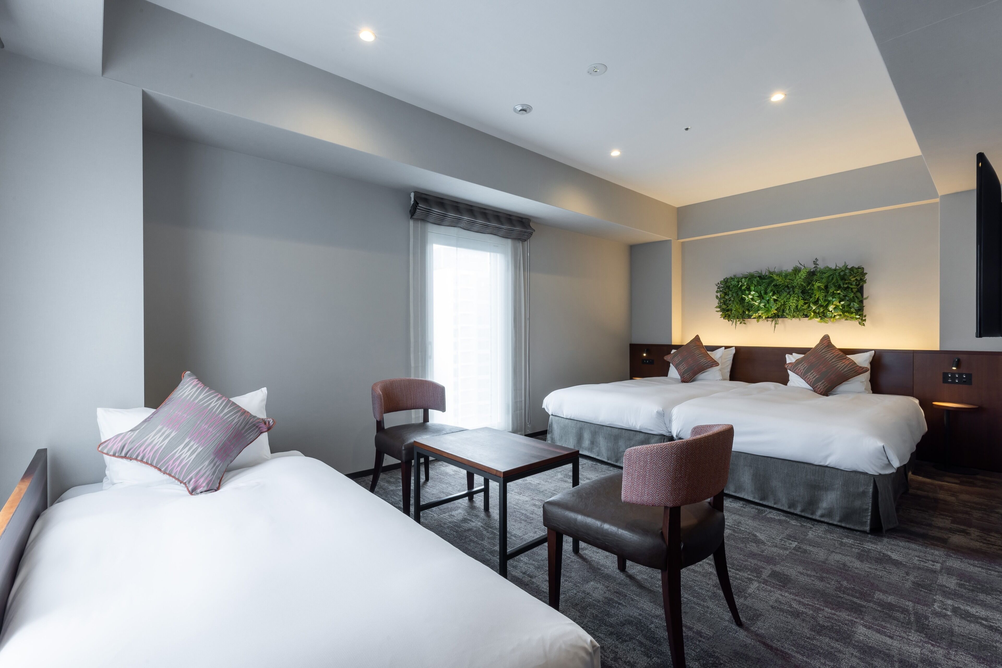 A hotel room with two beds, a small table, two chairs, a window with curtains, and a wall-mounted plant.