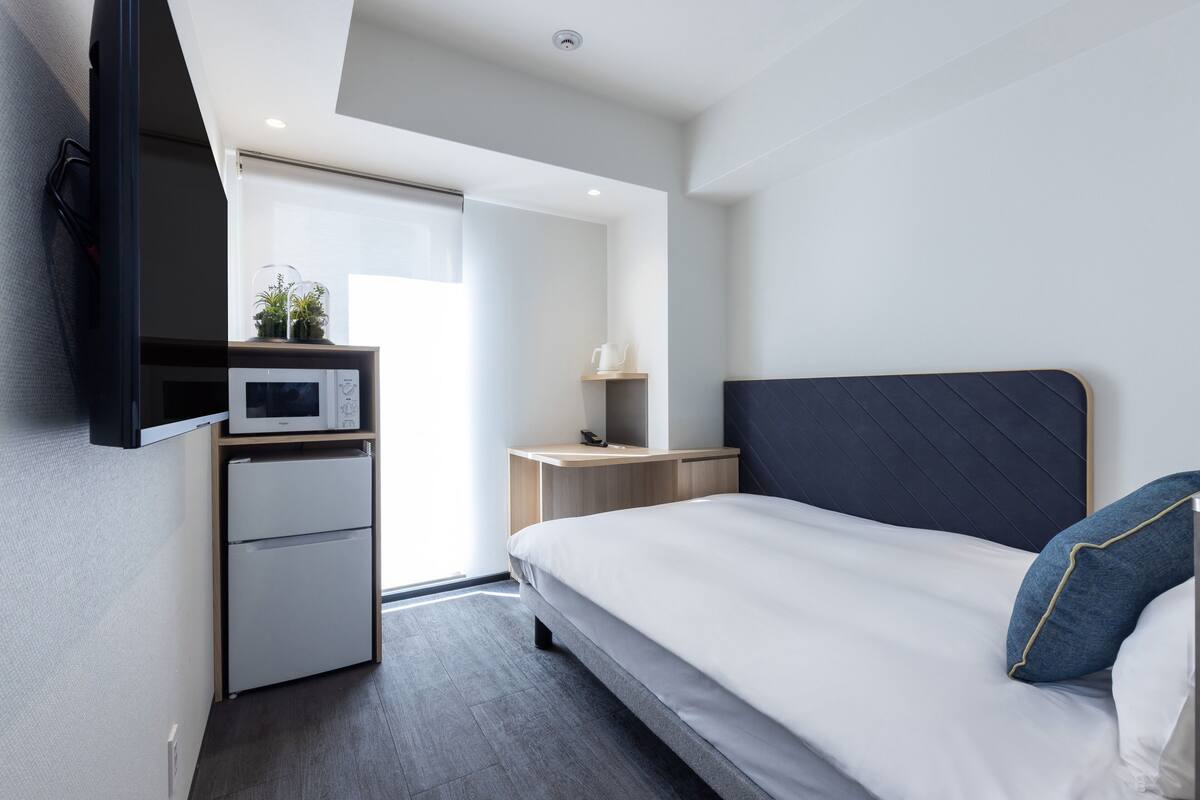 Ein modernes Hotelzimmer mit einem Bett, einem kleinen Schreibtisch mit einer Mikrowelle, einem Kühlschrank und einem Nachttisch mit einer Lampe.