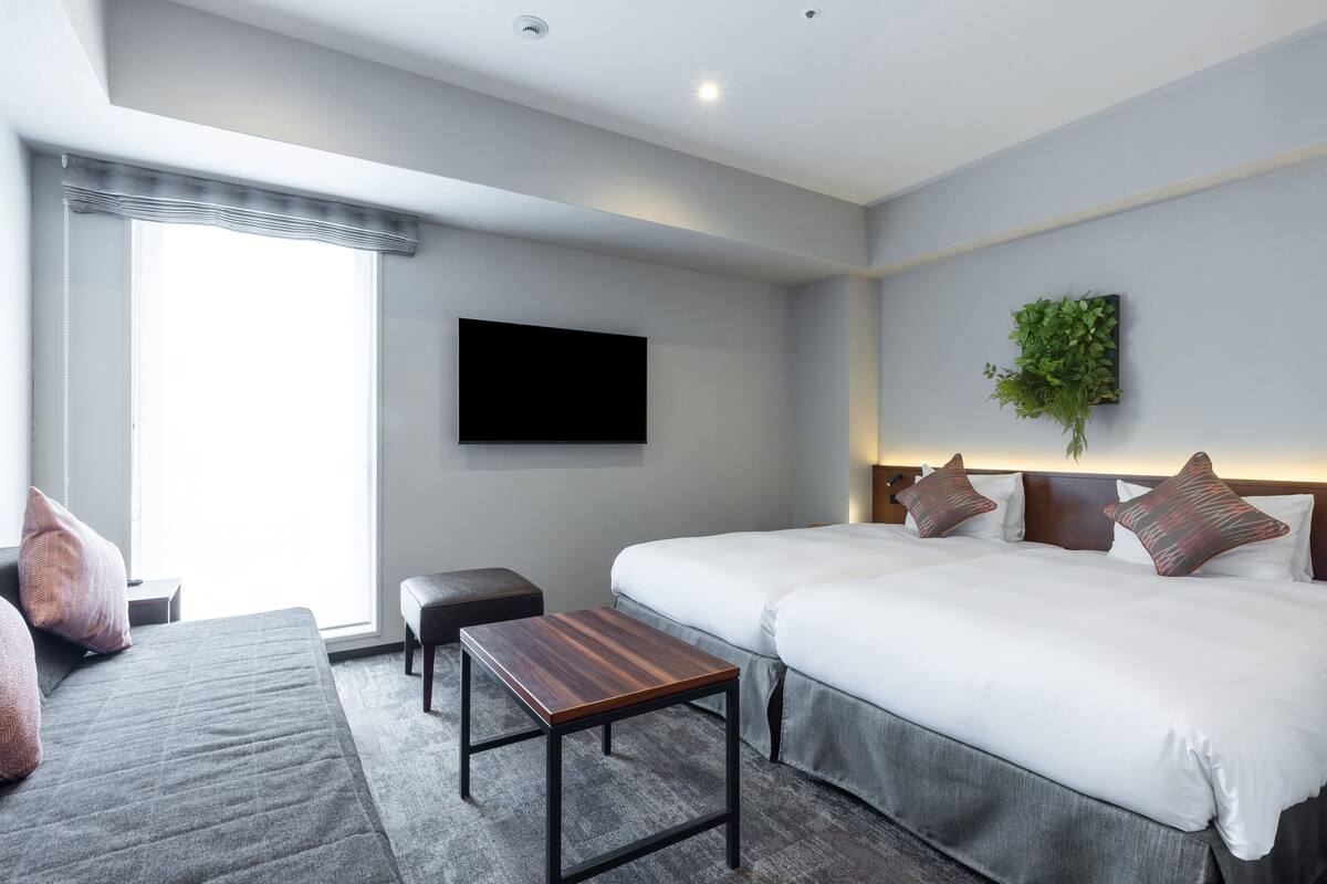 Ein Hotelzimmer mit zwei Betten, einem Flachbildfernseher, einem kleinen Tisch, einer Bank und einer Pflanze.