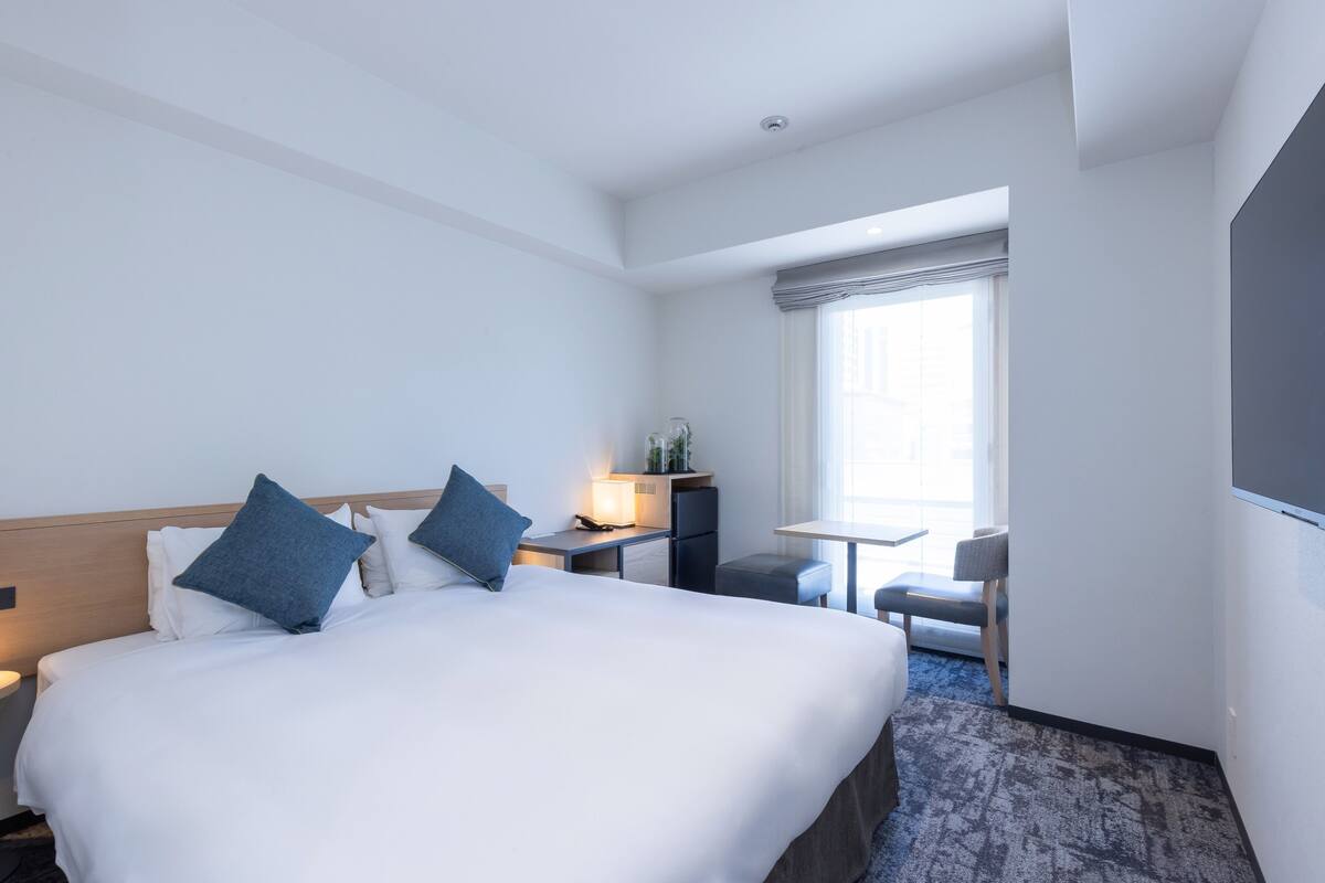 Ein Hotelzimmer mit einem großen Bett, einem Schreibtisch mit Stuhl, einem kleinen Tisch und einem Fenster mit Vorhängen.