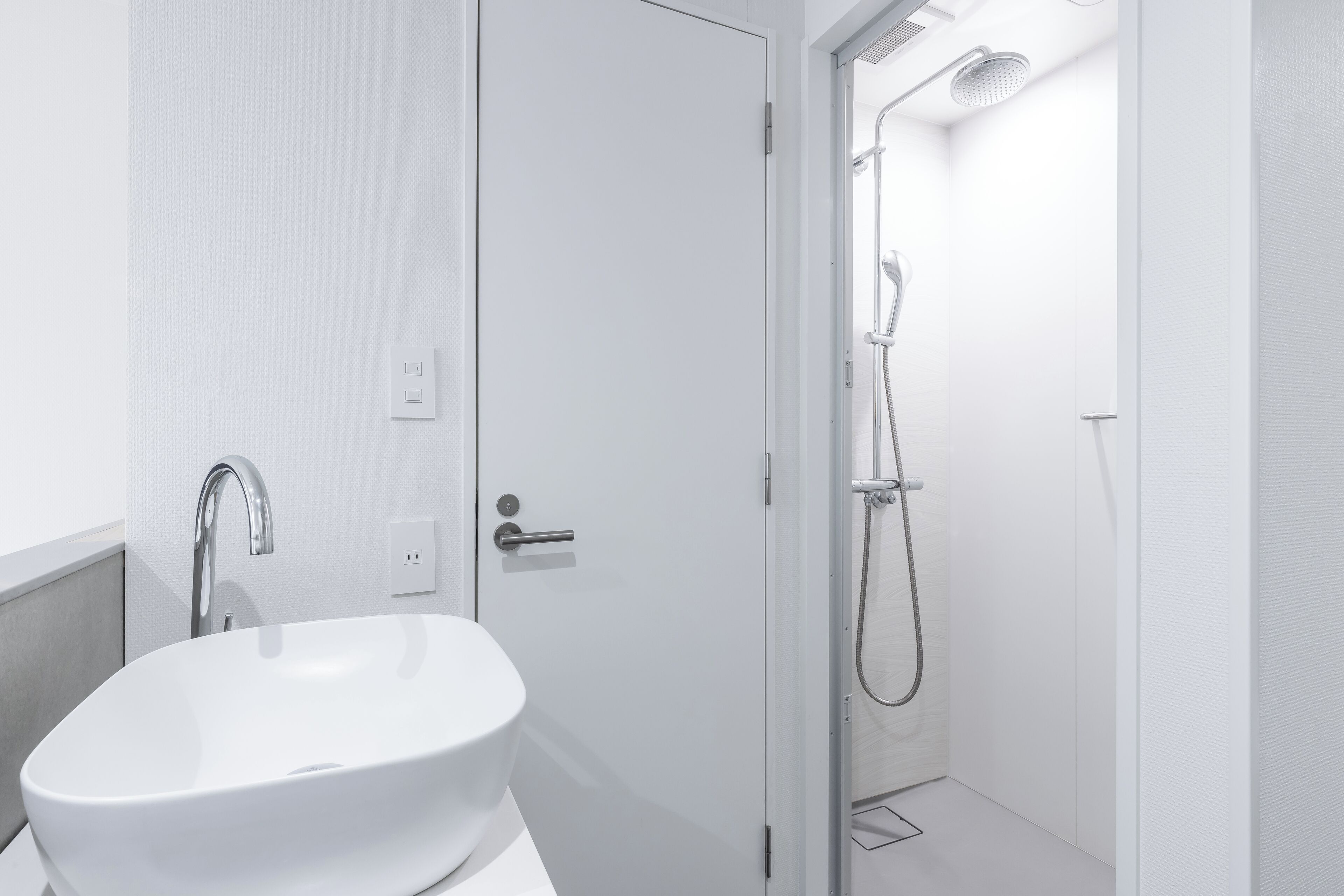 A modern bathroom with a white sink, a shower with a handheld showerhead, and a door leading to another room.