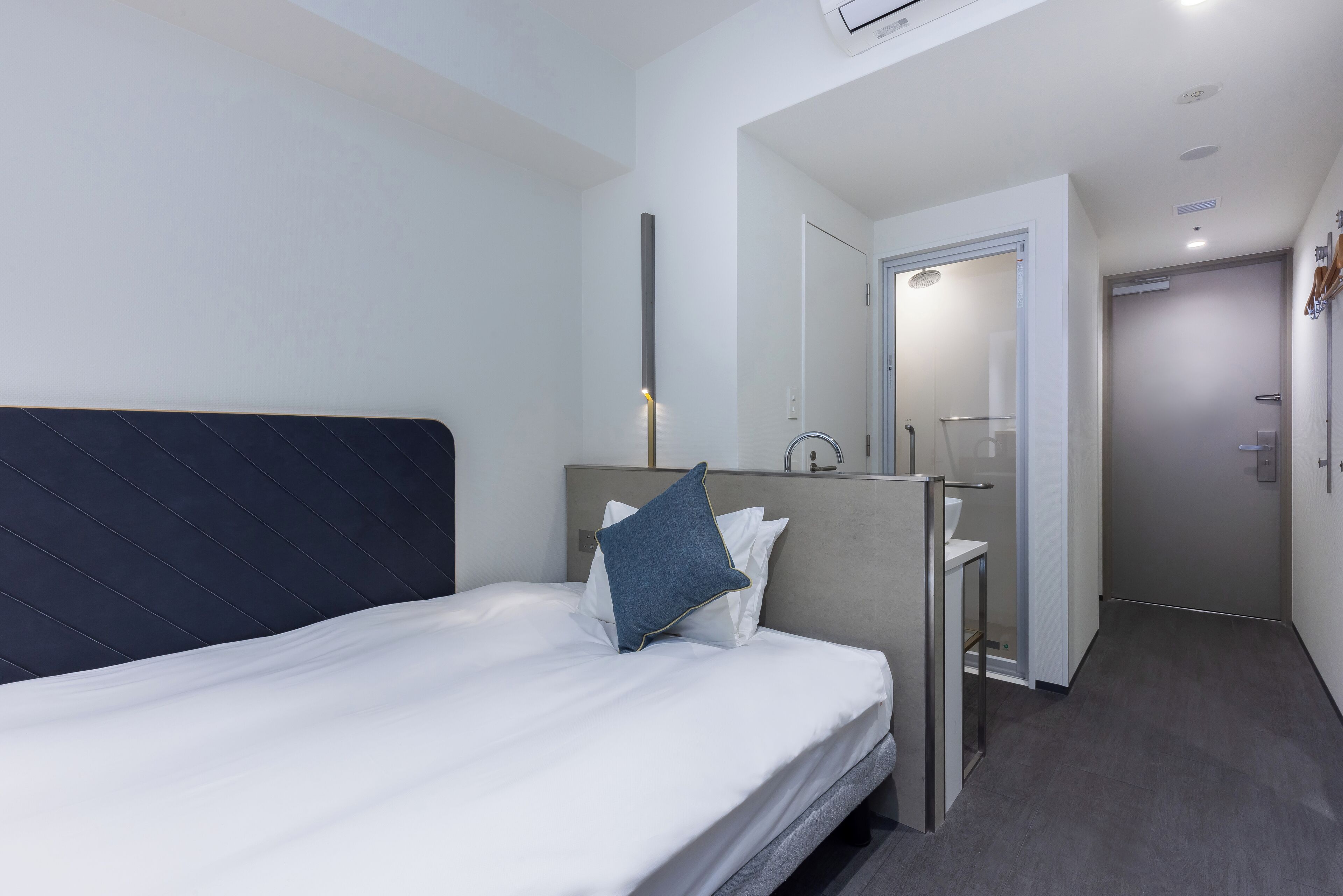A modern hotel room with a neatly made bed, a small bedside table, and a bathroom visible through an open door.
