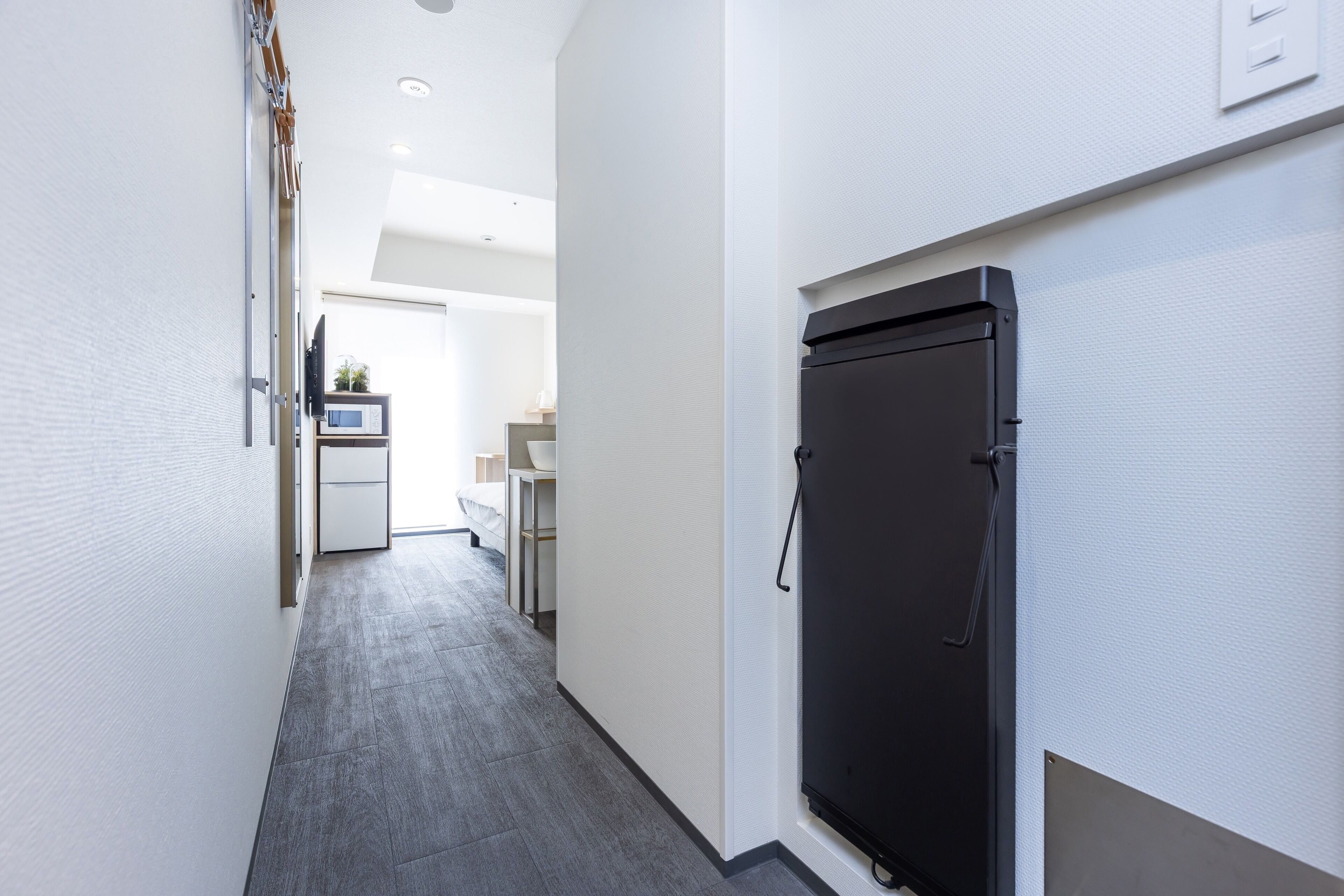A narrow hallway with a black refrigerator on the right, a white door at the end, and a small kitchenette area with a microwave and sink visible in the background.