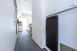 Um corredor estreito com uma geladeira preta à direita, uma porta branca no final e uma pequena área de cozinha americana com micro-ondas e pia visíveis ao fundo.