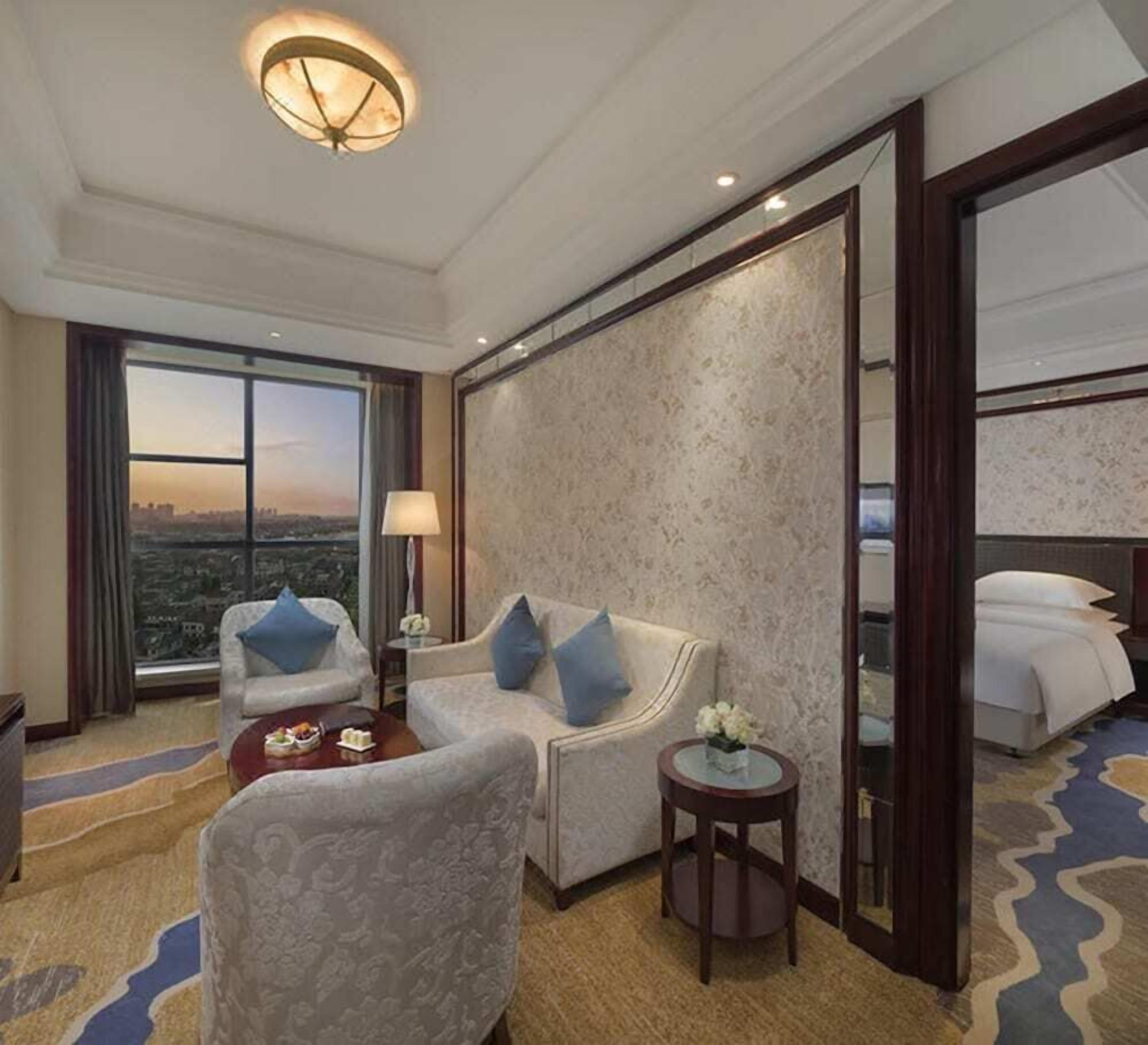 A hotel room with a large bed, two sofas, a coffee table, a small side table, a lamp, and a view of the city through a large window.