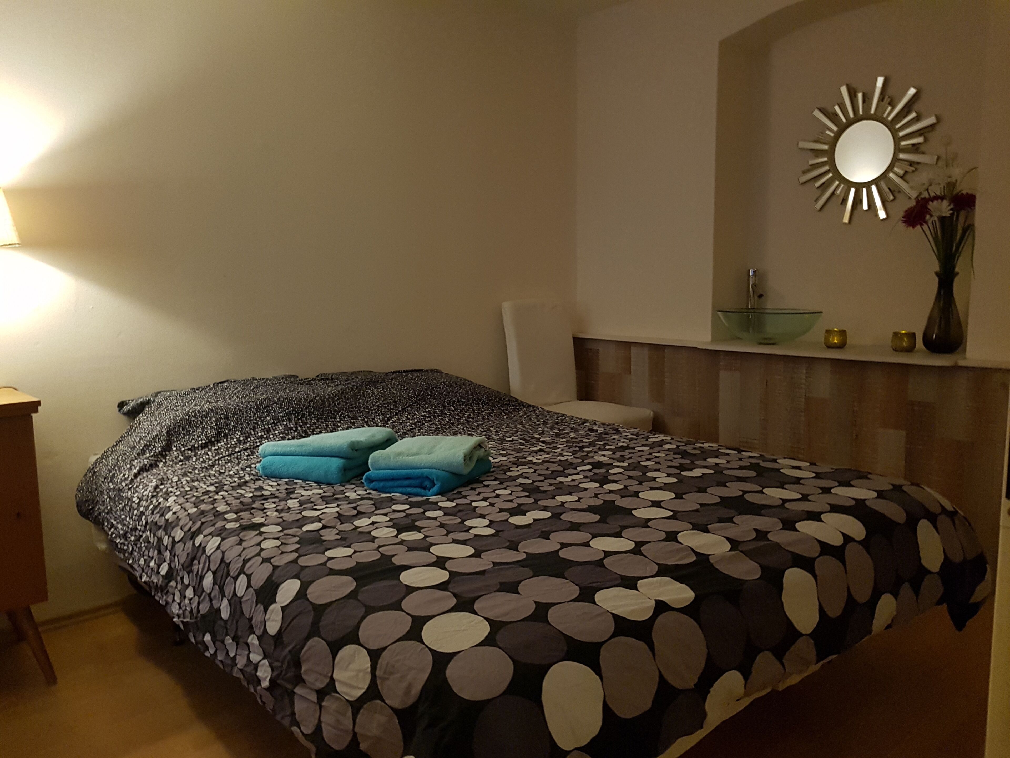 A bedroom with a bed, a nightstand, a mirror, and a vase with flowers.