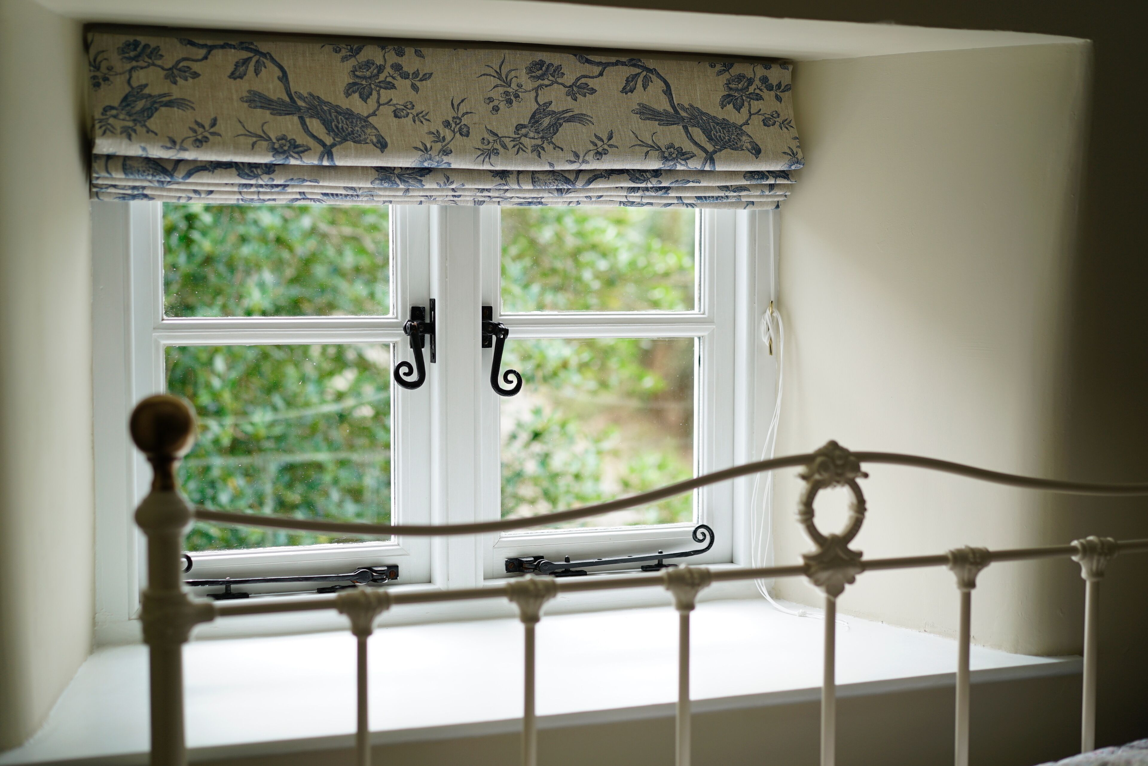 Una ventana con marco blanco y un adornado floral, una estructura de cama metálica blanca y vistas de vegetación afuera.