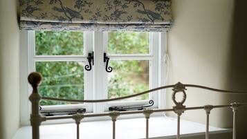 Una ventana con marco blanco y un adornado floral, una estructura de cama metálica blanca y vistas de vegetación afuera.