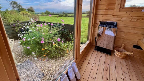 A wooden cabin with a large window offering a view of a garden with pink flowers and a clear sky.