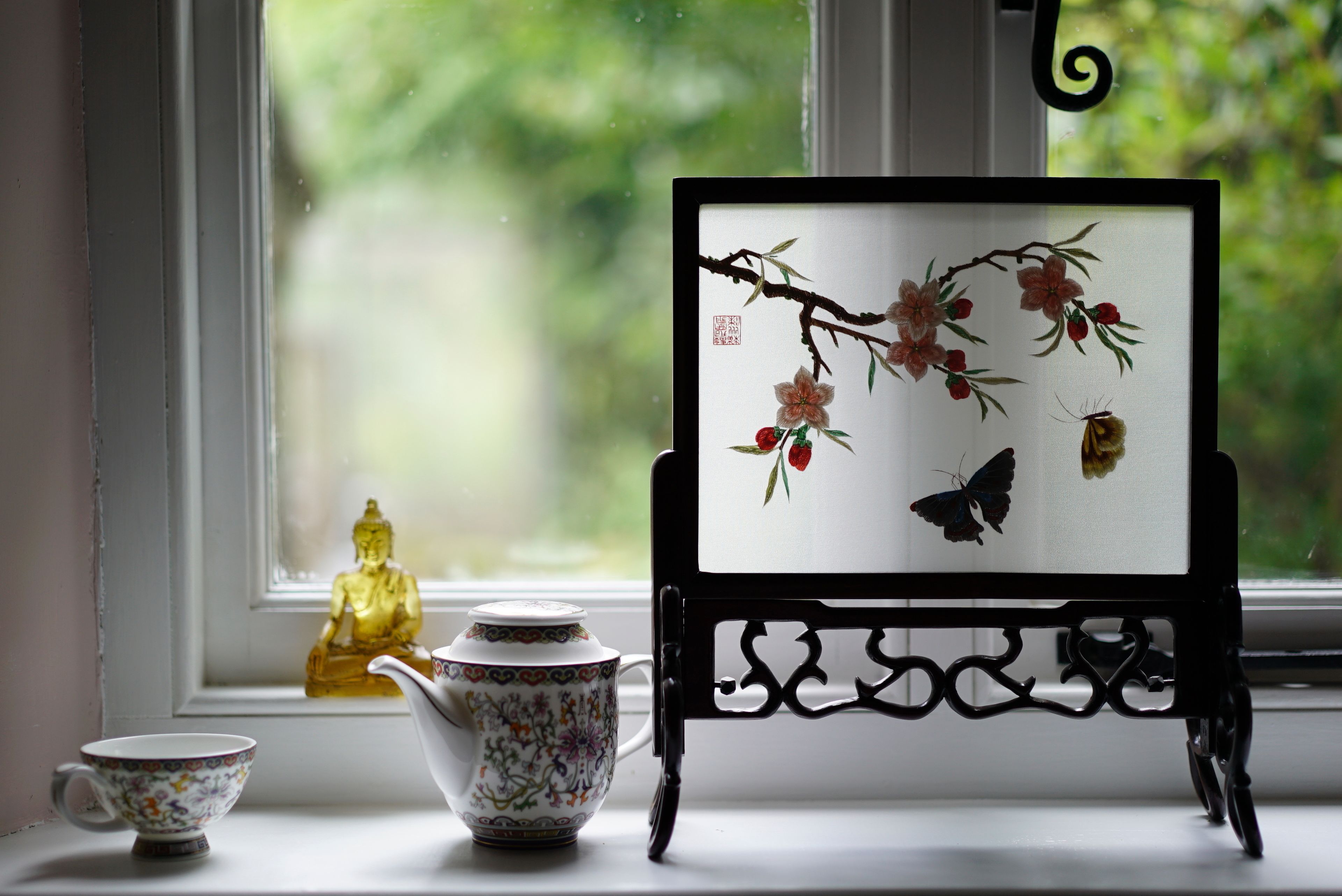 Una obra de arte en marco con flores y una mariposa, sobre un soporte junto a una ventana.