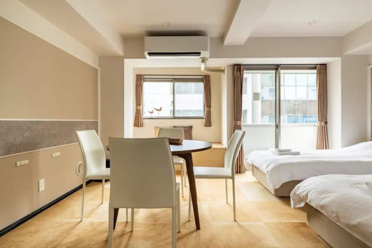 A hotel room with a bed, a dining table with chairs, a window with curtains, and an air conditioning unit.