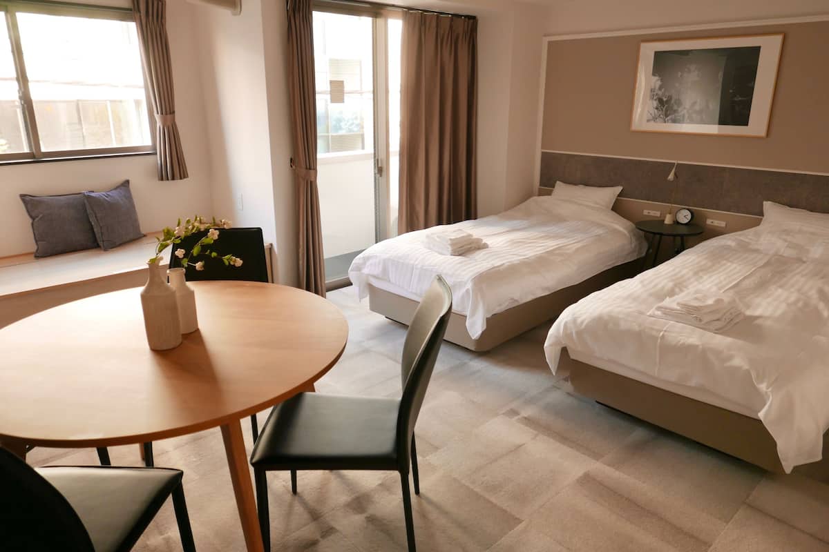 A hotel room with two beds, a dining table with chairs, a small table with a clock, and a window with curtains.