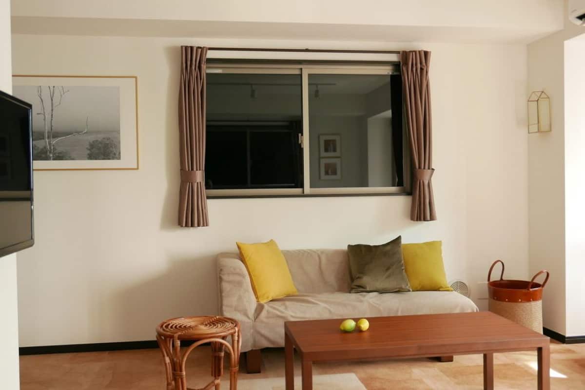 A living room with a sofa, a wooden table, a wicker chair, a window with curtains, and a framed picture on the wall.