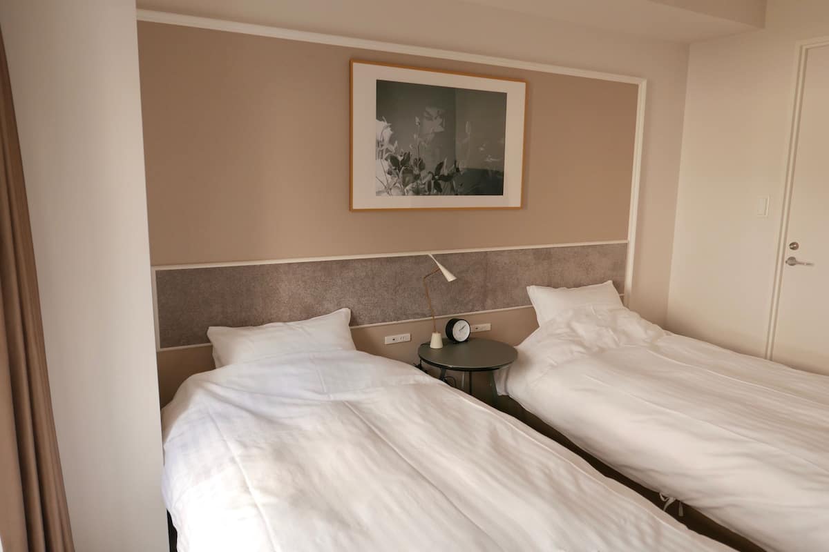 A hotel room with two single beds, a bedside table with a clock, and a framed picture on the wall.