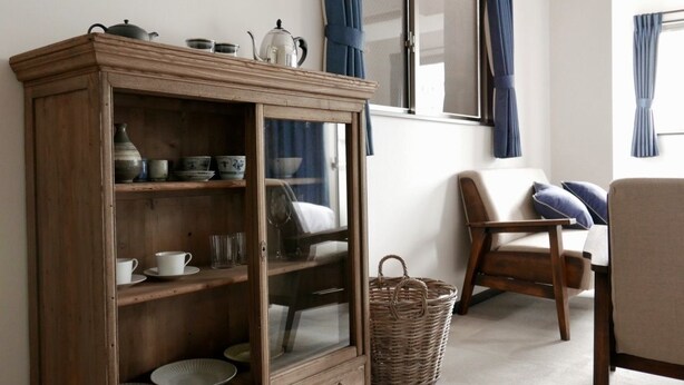 A wooden cabinet with glass doors, a wicker basket, a wooden chair, and a sofa with blue cushions in a room with a window and curtains. - Tokyo Sakurako Hatchobori (Tokyo)