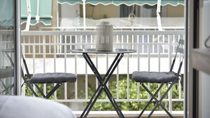 A balcony with a table and two cushioned chairs, a metal container, and a view of a building with a green awning. - Live As A Greek in Artemis Apartment (Athens)
