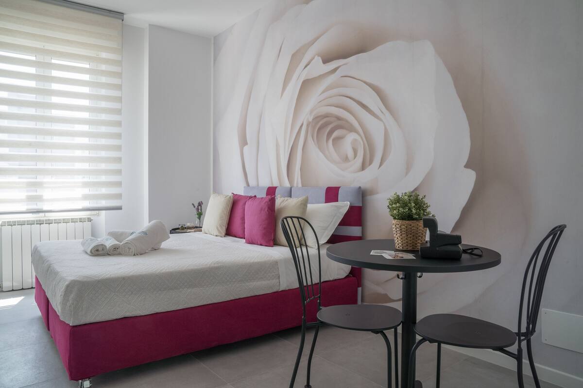 Un dormitorio con un amplio mural en la pared, una cama con muchas almohadas, una pequeña mesa redonda con sillas y una planta en maceta.