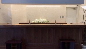 A reception area with a dark wooden counter, a floral arrangement, and two small tables. - Yamanashi Grand Hotel (Chuo)