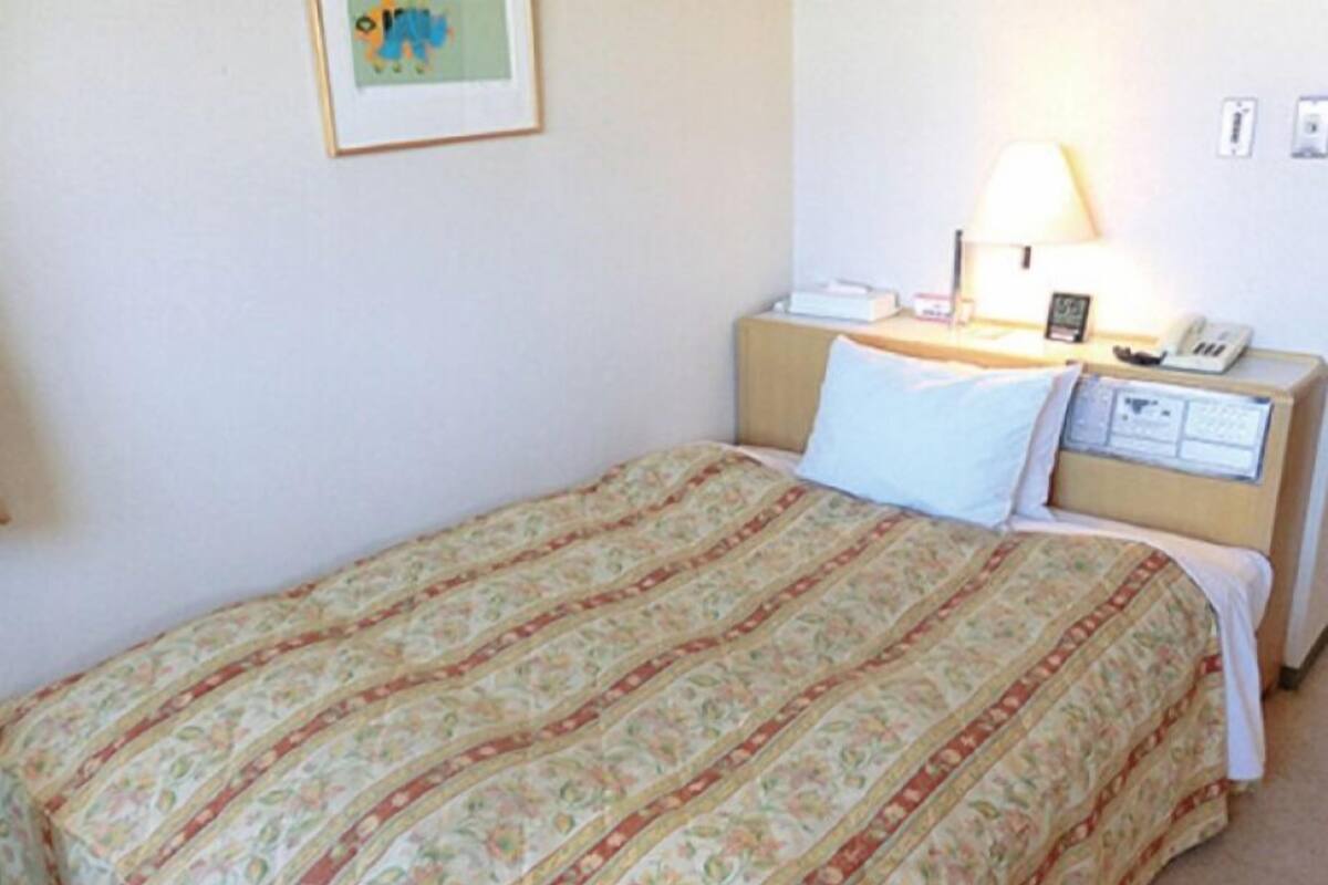 A single bed with a floral-patterned bedspread, a bedside lamp, a nightstand with a telephone, and a framed picture on the wall.