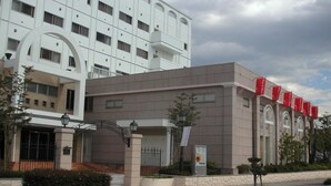 A modern building with a beige facade, a large arched entrance, and red banners on the right side. - Yamanashi Grand Hotel (Chuo)