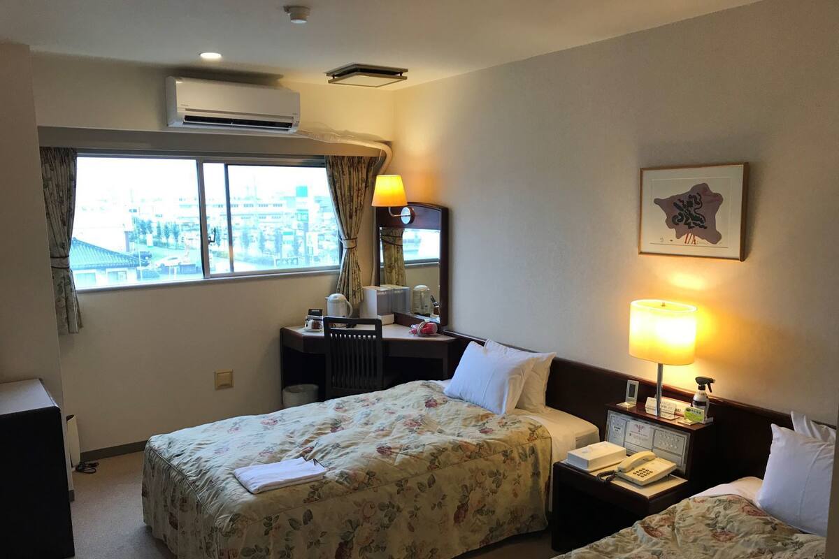 A hotel room with two beds, a desk, a chair, a lamp, a telephone, and a window with curtains.