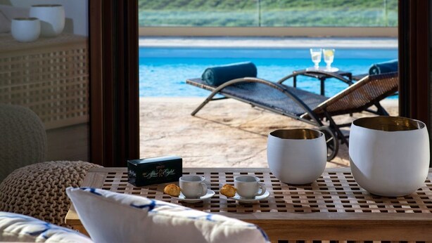 A room with a view of the beach, a table with cups and a box of tea, and a chair by the window.