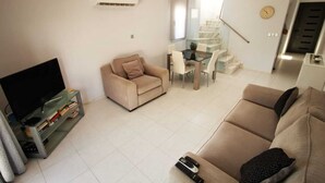 Ein Wohnzimmer mit einer Couch, einem Fernseher, einem Esstisch mit Stühlen, einer Treppe und einer Klimaanlage.