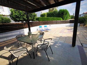 A covered patio area with a glass table and wicker chairs, a blue and white striped lounge chair, and a bench. - Sea side holiday villa, perfect location, maximum relaxation. (Chlorakas)