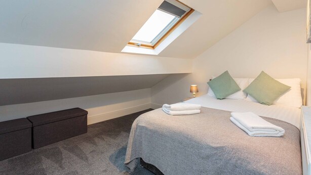 A bedroom with a skylight, a bed with pillows, a bedside table, and a storage bench. - Cherry Property - Apple Suite (Blackpool)