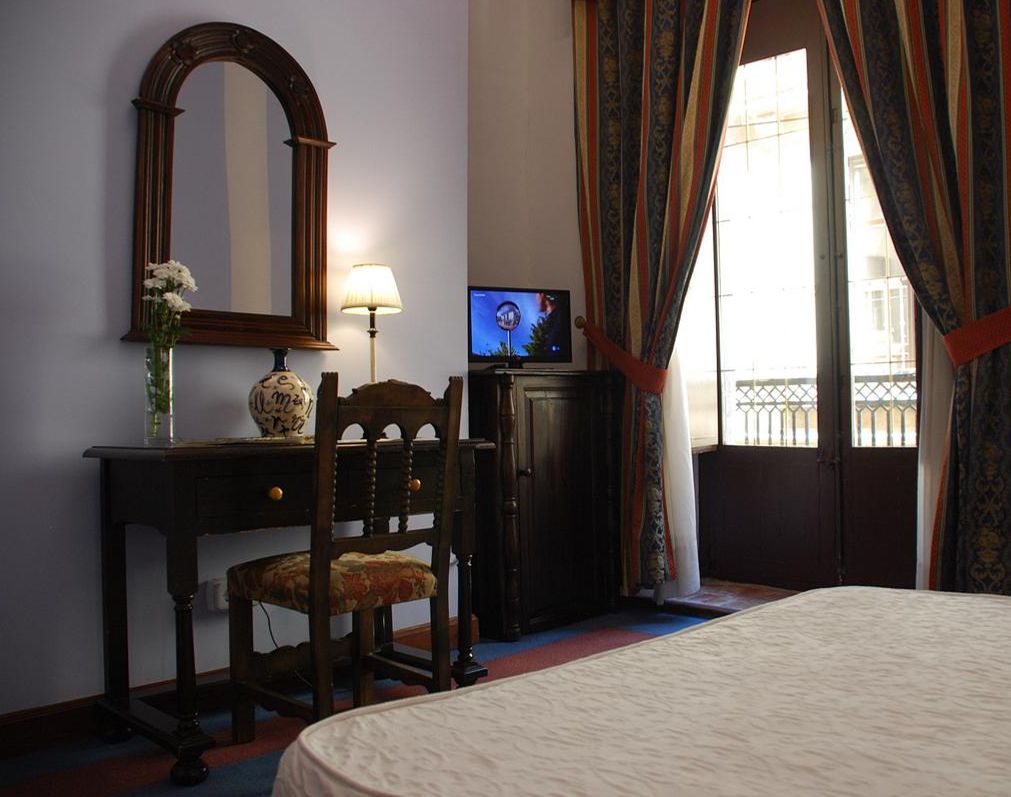 Hotelkamer met een bed, een bureau met lamp, een televisie, een spiegel en een raam met gordijnen.