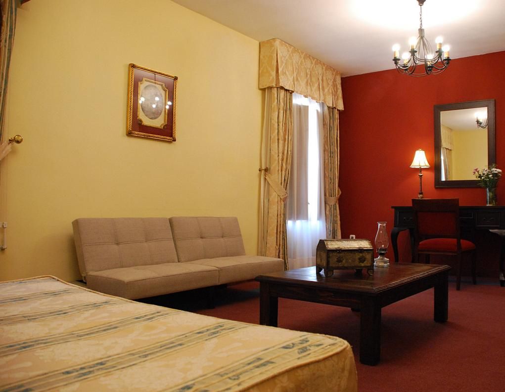 A hotel room with a sofa, a small table, a bed, a chandelier, a mirror, and a window with curtains.
