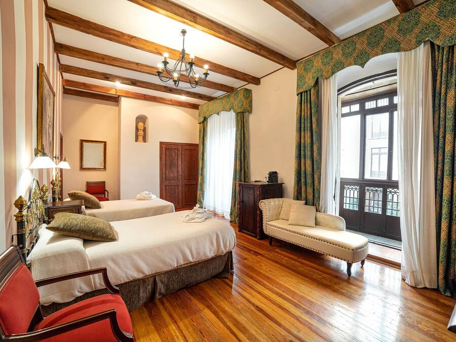A hotel room with two beds, a sofa, a chair, a wooden floor, a chandelier, and a large window with curtains.