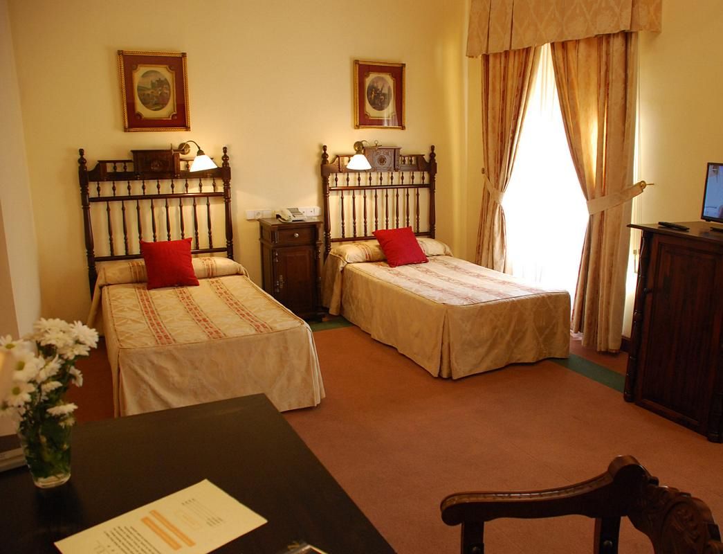 A hotel room with two single beds, a desk with a chair, a small table with a vase of flowers, and a desk lamp.