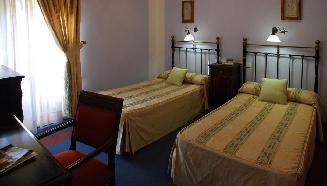 A room with two single beds, a desk with a chair, and a window with curtains.
