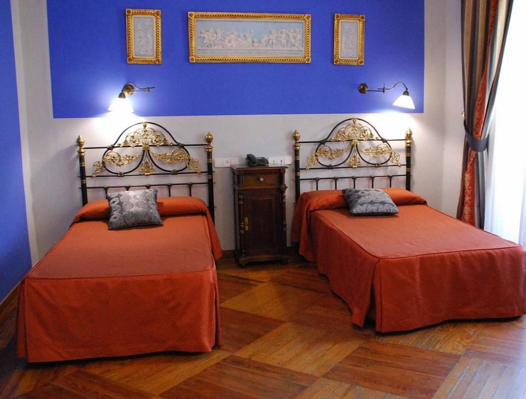 Una habitación con dos camas, cada una con una colcha roja y un cojín a juego. Hay lámparas fijadas a la pared y cuadros enmarcados sobre las camas.