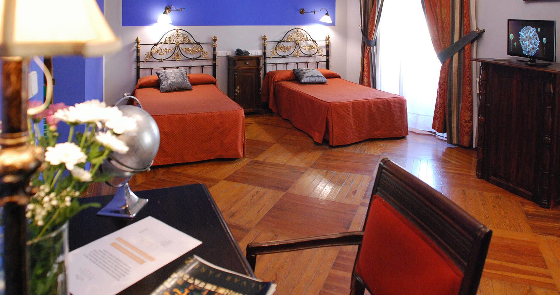 A hotel room with two single beds, a wooden floor, a desk with a lamp, and a television.